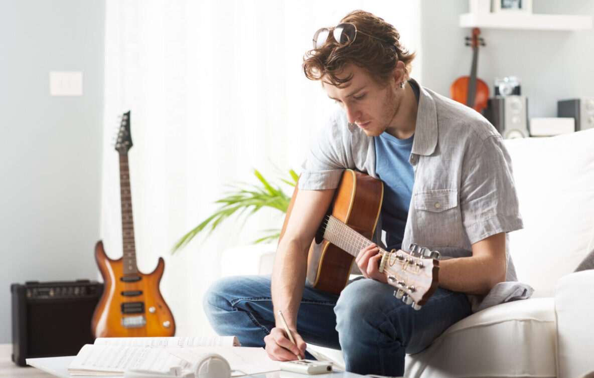 A college student is sitting with an acoustic guitar in his arms. He is writing music. An electric guitar is pictured in the background.