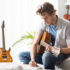 A college student is sitting with an acoustic guitar in his arms. He is writing music. An electric guitar is pictured in the background.