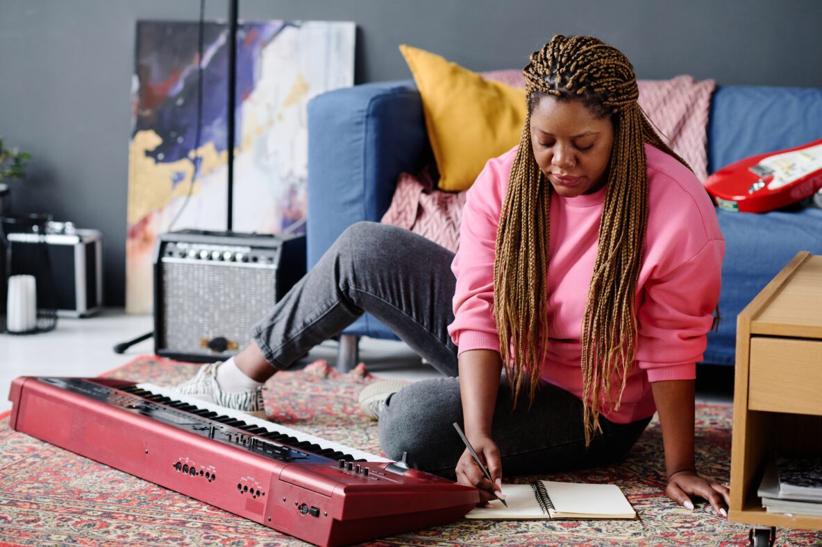 A young woman is sitting on the floor in front of a keyboard. She is writing on a piece of paper. Behind her is an amp and electric guitar.