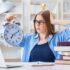 A stressed looking college student is looking at and holding a giant analog clock in one hand.
